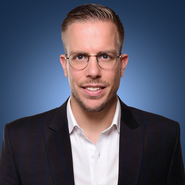 Person smiling, wearing glasses and a suit, against a blue background.