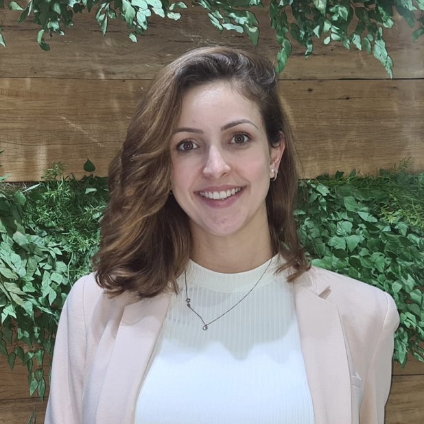 Person smiling, standing in front of a wooden wall with green plants.