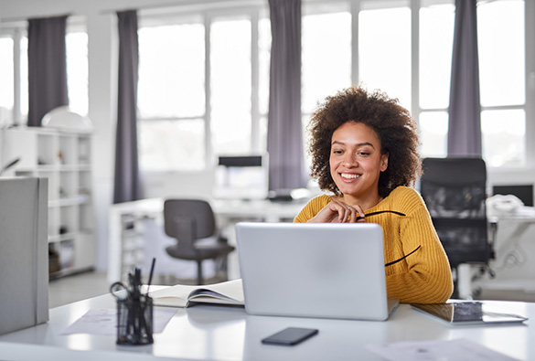 Person smiling while using a laptop in a bright office.