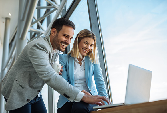 Two people smiling and looking at a laptop by a large window.