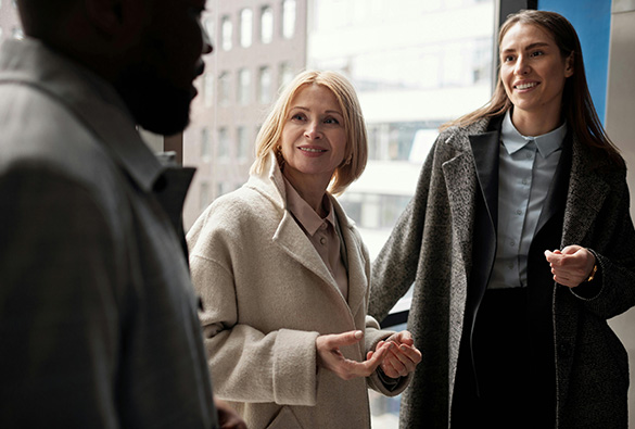 Three people in conversation by a window wearing coats.