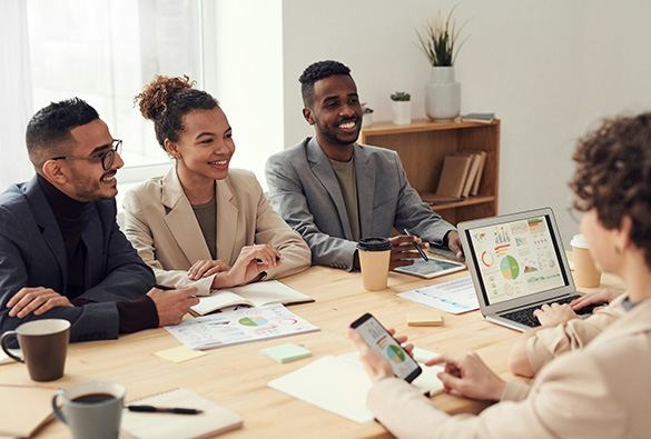 A group of people in a meeting room discussing charts displayed on a laptop with coffee cups on the table.