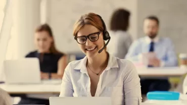 Person wearing glasses and headset, smiling while working on a laptop in an office.