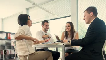 Group of colleagues in a meeting at an office table, laptops and files visible.