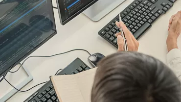Person writing notes while discussing code on computer screens.