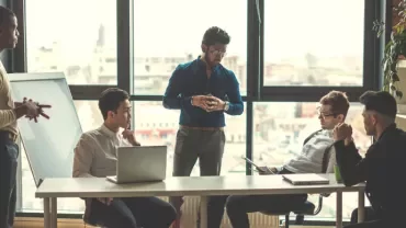 A team meeting in a bright office with five people discussing around a table.