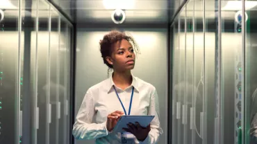 Person working in a server room, holding a tablet and observing data panels.