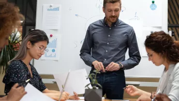 Group of people in a meeting room discussing with a whiteboard in the background.