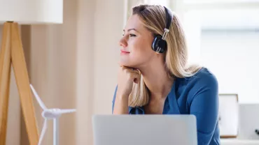 Person with headphones looking out the window, seated at a desk with a laptop.
