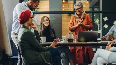 A diverse group of people in a casual office meeting, laughing and smiling around a table with a laptop.