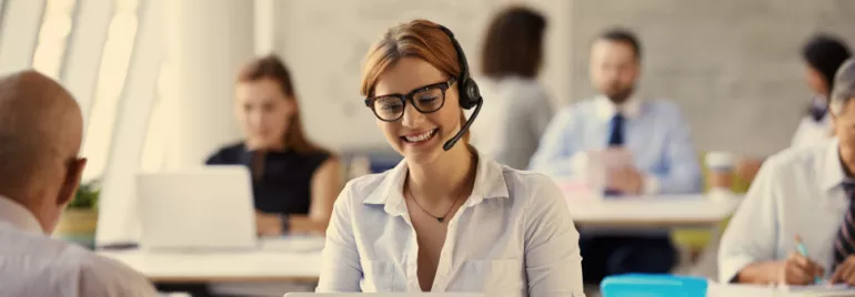 Person wearing glasses and headset, smiling while working on a laptop in an office.