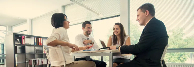 Group of colleagues in a meeting at an office table, laptops and files visible.