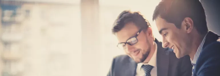 Two people in suits smiling and looking at a laptop in a bright office.