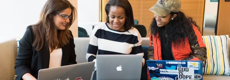 Three people sitting on a couch, working on laptops with various stickers.