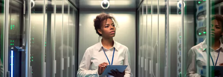 Person working in a server room, holding a tablet and observing data panels.