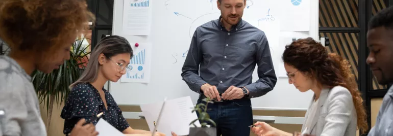 Group of people in a meeting room discussing with a whiteboard in the background.