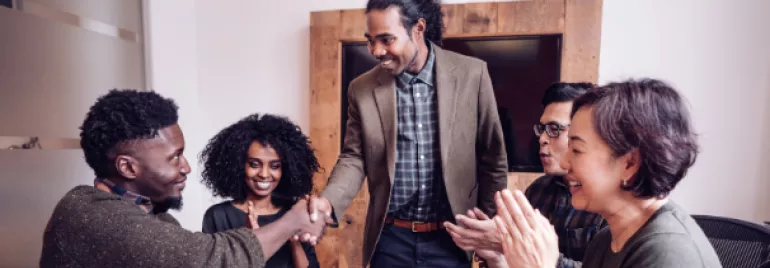 Smiling colleagues clapping as two people shake hands in an office.