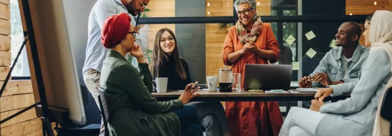 A diverse group of people in a casual office meeting, laughing and smiling around a table with a laptop.