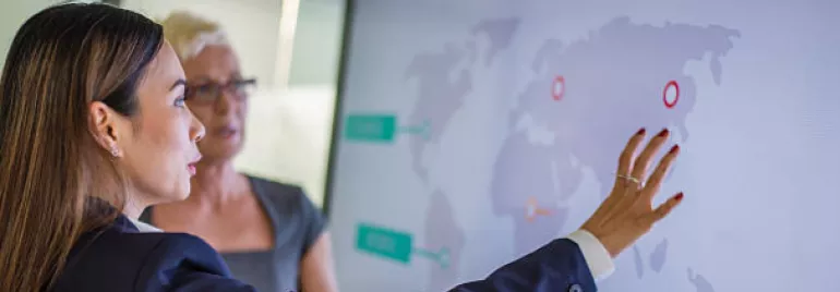 Two people discussing data displayed on a large screen with a world map.