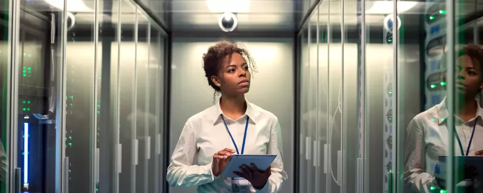 Person working in a server room, holding a tablet and observing data panels.