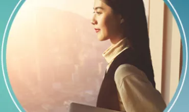 Person with a laptop looking out a window at a cityscape during sunset.