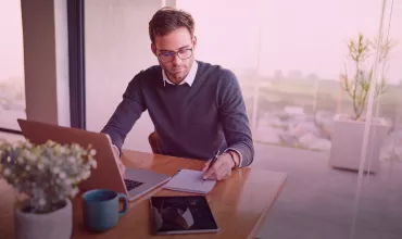 Person writing notes at a desk with a laptop, notebook, and tablet in a bright room.