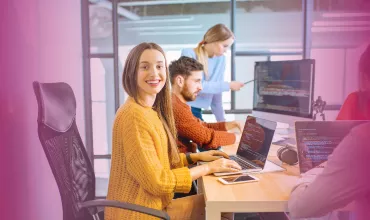 Person smiling at a desk with open laptop, others working on computers in a modern office.
