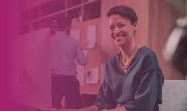 Person smiling at a desk with a colleague in the background.
