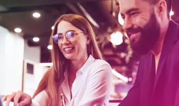 Two people smiling while working on a laptop in a brightly lit office.