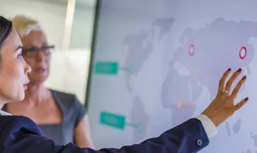 Two people discussing data displayed on a large screen with a world map.
